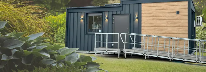 Außenansicht des TinyCareHome Modernes TinyCareHome-Modulgebäude mit barrierefreier Rampe in parkähnlicher Umgebung. Das kompakte Gebäude kombiniert dunkle Metallfassade mit Holzverkleidung und verfügt über eine Eingangstür mit Beleuchtung.